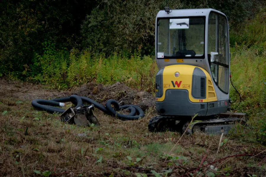 Compact excavator in long grass beside coils of black drainage pipe