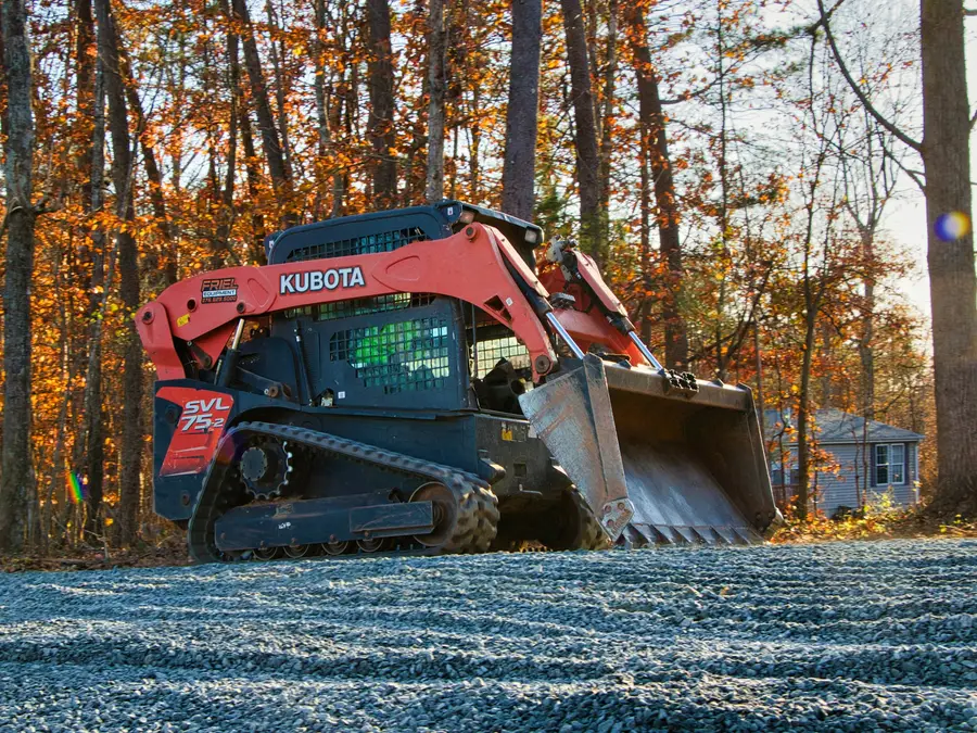 Compact track loader spreading gravel on a driveway among autumn trees