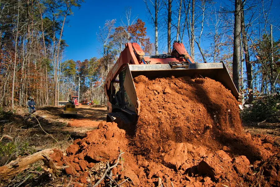 Compact track loader lifting a bucket of red soil on a wooded lot