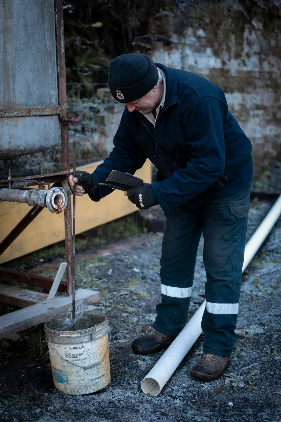 Worker in gloves and a toque handling a length of white pipe beside a bucket